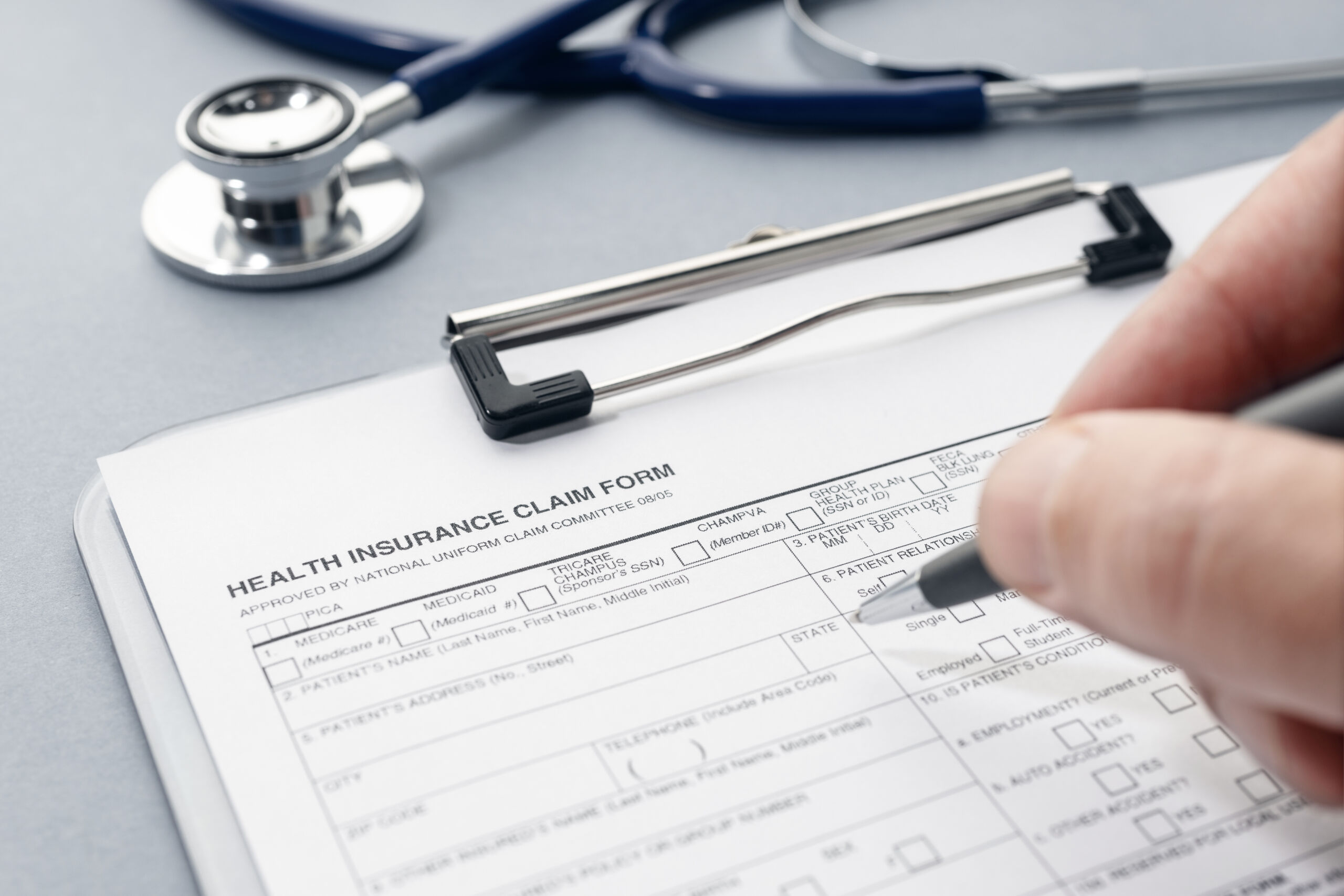 Insurance Accepted & Authorization Help | Jubilee Speech Therapy LLC | Daphne, AL Hand filling out a health insurance claim form on a clipboard with a stethoscope in the background, representing insurance verification and approvals for therapy services.